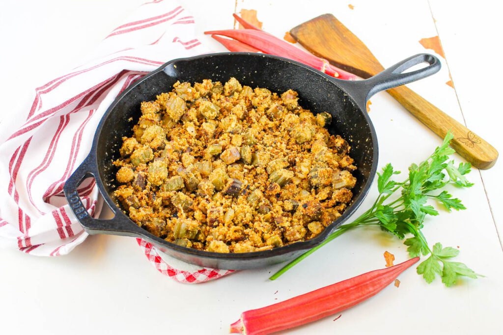 cast iron skillet sits next to a striped tea towel, fresh picked okra and some green herbs