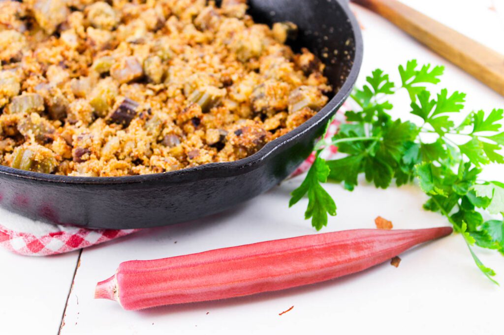 a fresh pod of okra sits in front of a pile of herbs and a skillet with fried food