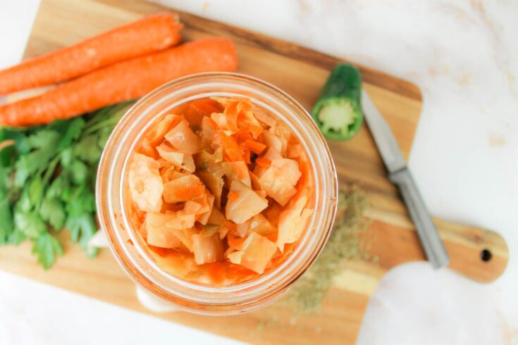 jar of fermented curtido recipe sitting on a cutting board surrounded by fresh herbs and vegetables