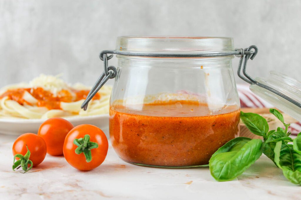 glass jar filled halfway with from scratch spaghetti sauce on a countertop covered with fresh herbs and cherry tomatoes