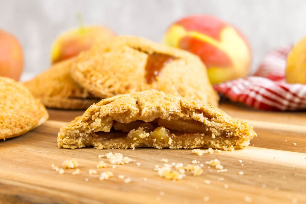 half of an apple hand pie with fresh milled flour sits on a wooden surface surrounded by apples