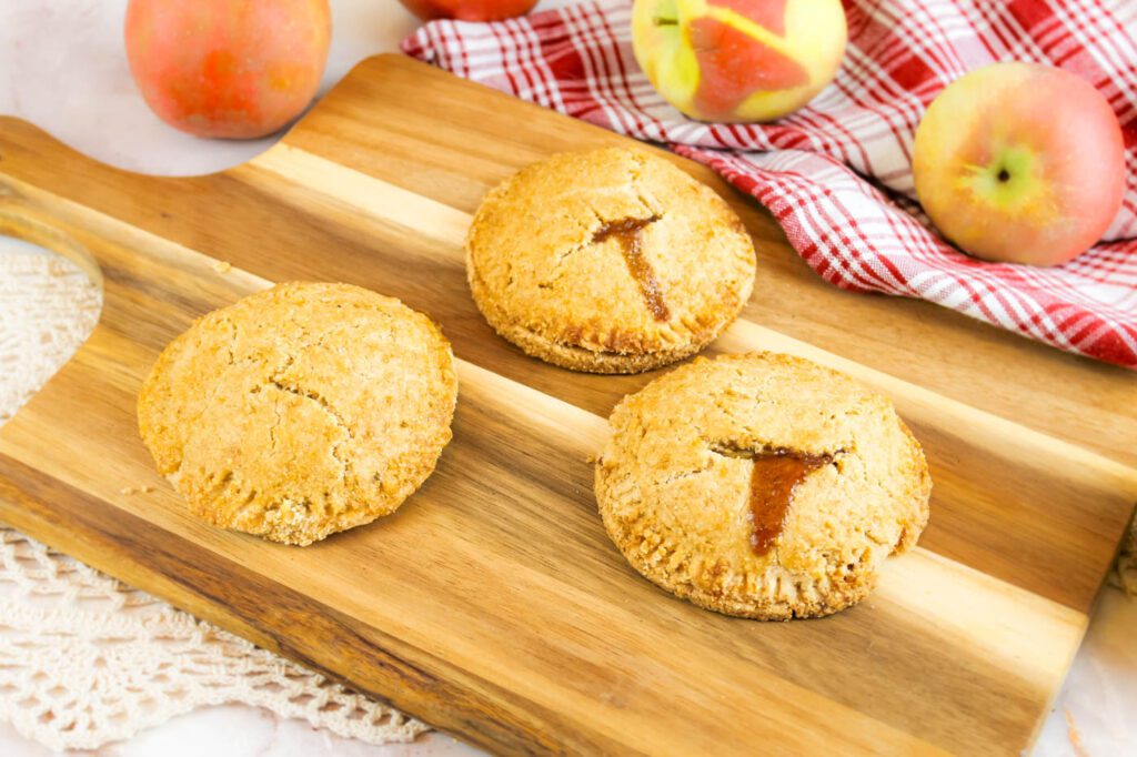 3 small apple pies on a cutting board besides a red plaid kitchen towel