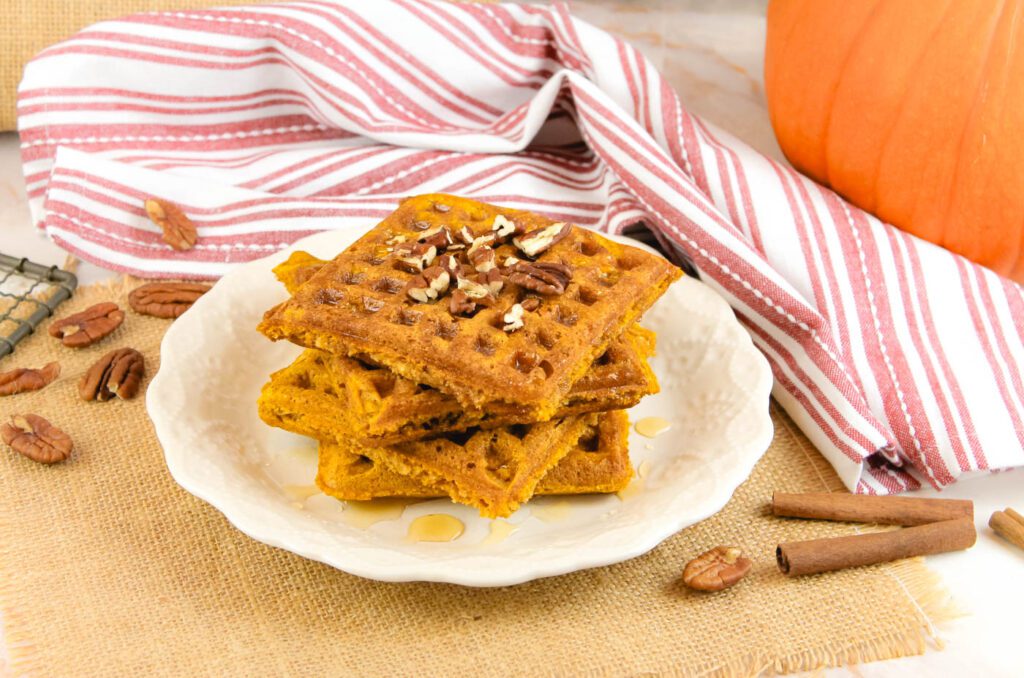 a white plate with a stack of three pumpkin waffles sits beside a pumpkin, cinnamon sticks and a pile of pecans