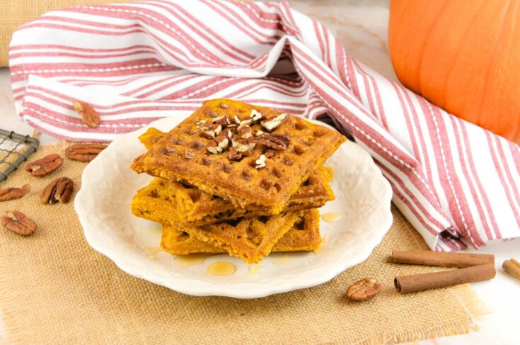 a white plate with a stack of three pumpkin waffles sits beside a pumpkin, cinnamon sticks and a pile of pecans