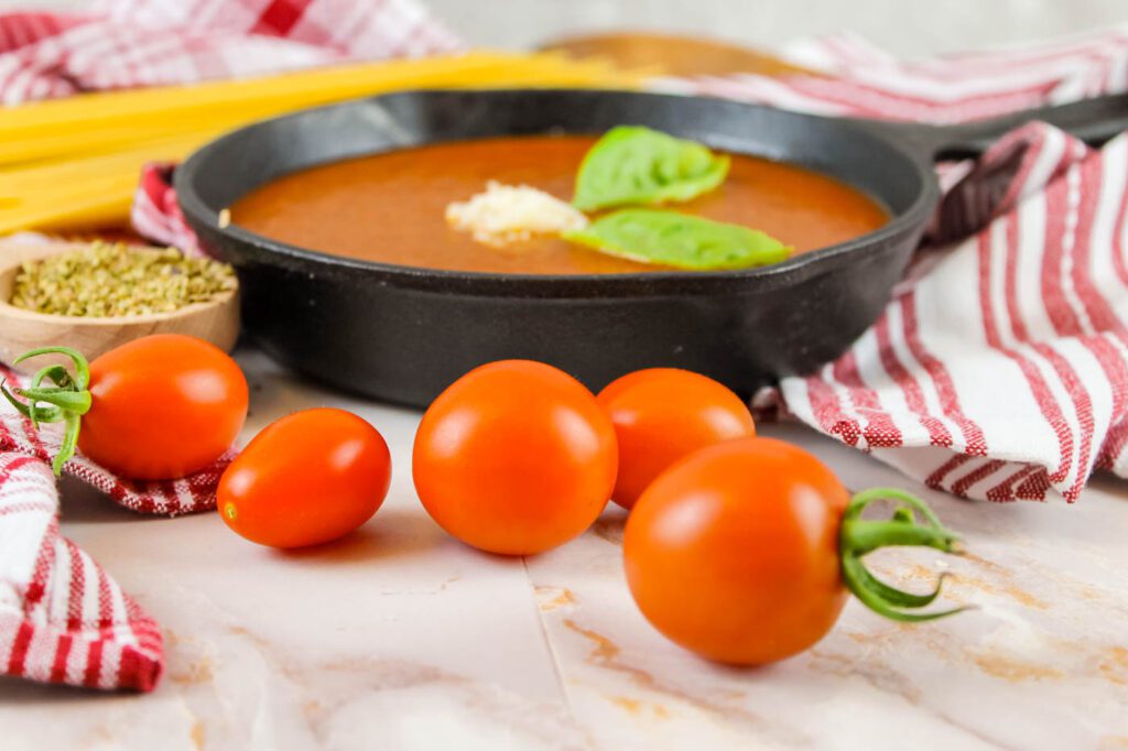 5 red tomatoes sit in front of a pan filled with marinara sauce