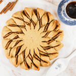 Cappuccino Cinnamon Star Bread sits beside a cup of coffee, coffee beans, a pie server and some cinnamon sticks