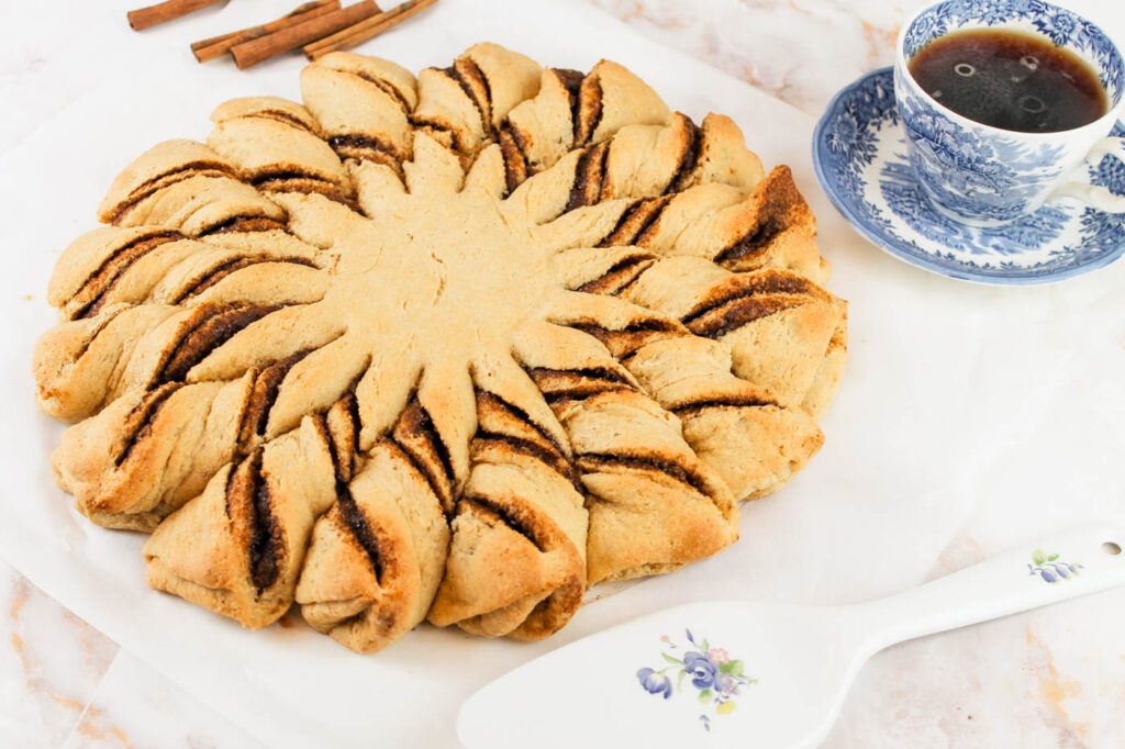 twisted cinnamon roll on a marble kitchen countertop sits beside a fresh cup of coffee