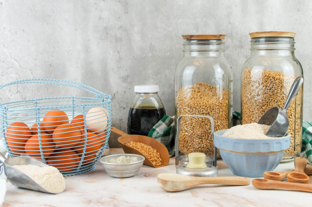 two glass jars filled with wheat berries, a basket of farm fresh eggs, a bottle of honey, a bowl of fresh milled flour and various baking tools sit on a marble countertop