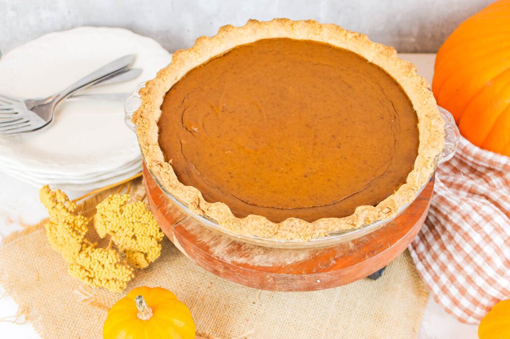 pie on a display stand next to a stack of plates, a pile of forks and some fall decor