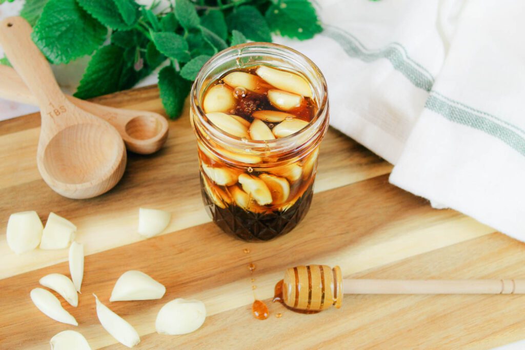 jar of fermented garlic and honey sits beside fresh garlic cloves, measuring spoons, a kitchen towel and a dipper covered in raw honey