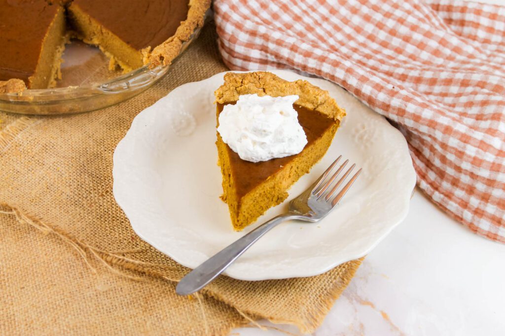 slice of pumpkin pie covered with cream sits beside a brown checkered towel and whole pie