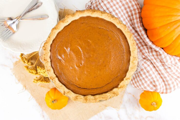 pumpkin pie with fresh milled pie crust sits on a table beside plates, forks and fresh pumpkins