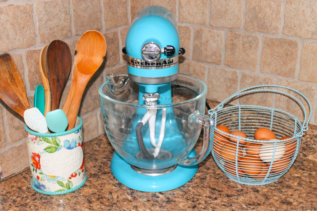 a kitchen aid mixer on a counter beside a jar of wooden spoons and a basket of eggs