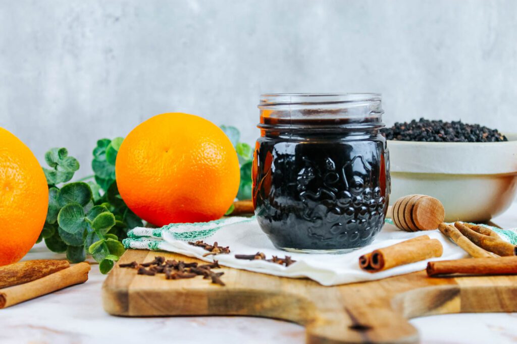 glass jar of herbal syrup beside whole spices and fresh oranges.