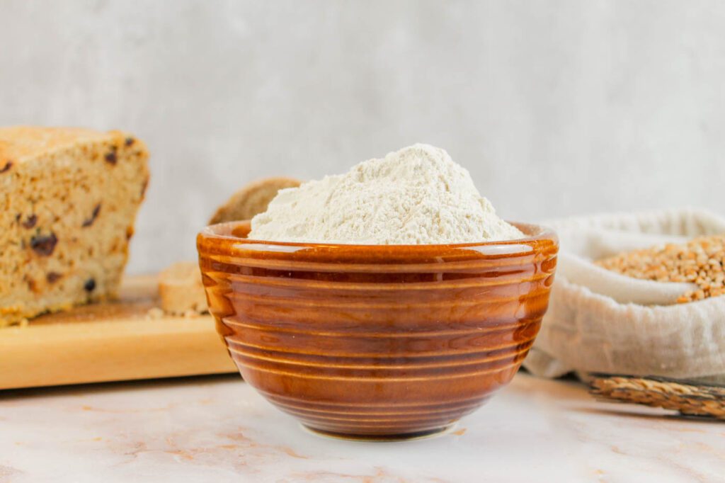 bowl filled with powdered gluten sits in front of a tray of bread and whole grains