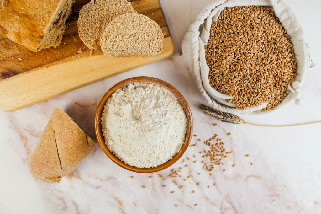 a top view of a kitchen counter with a bowl of gluten, two loaves of bread and a bag filled with wheat berries