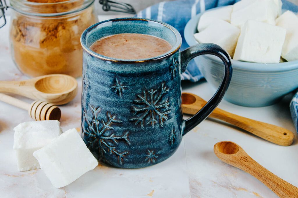 ceramic mug sits beside homemade marshmallows, wooden spoons and a glass container of cocoa powder
