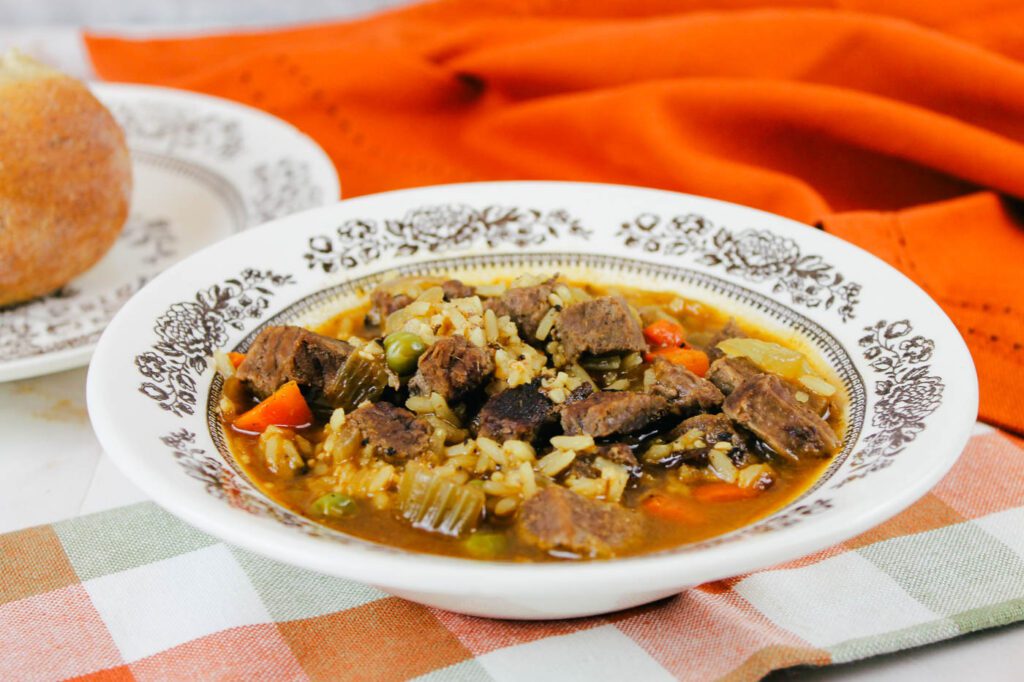 a brown and white floral bowl filled with beef soup sits next to some kitchen towels and a homemade roll