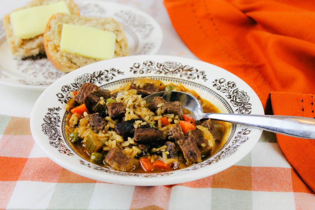 brown floral bowl filled with beef rice soup sitting beside an orange napkin and a plate of rolls covered in butter